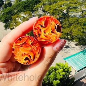May include: A pair of orange and yellow swirl acrylic ear gauges. The gauges are held in a hand with a painted fingernail.
