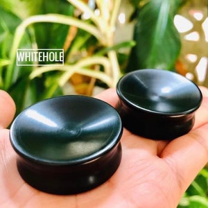 May include: Two black wooden ear gauges, one larger than the other, with a smooth, slightly concave surface. The gauges are held in a hand.