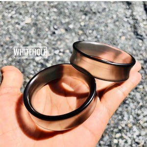 Può includere: Due dilatatori per orecchie grigio scuro sono tenuti in una mano. I dilatatori sono circolari con un bordo svasato. Il testo "WHITEHOLE" è in un riquadro bianco in alto a sinistra.