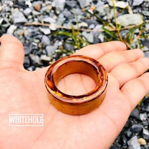 May include: A circular, amber-toned wooden ring is held in an open hand. The ring has a glossy finish and a lighter interior. The word "WHITEHOLE" is printed in a white rectangular box on the lower left side of the image.