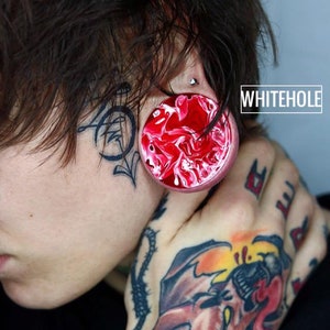 Puede incluir: Una persona con un expansor de oreja de acrílico rojo y blanco con forma de remolino, un tatuaje en la cara y un tatuaje de un murciélago en la mano. El expansor de oreja es de gran tamaño y está hecho de acrílico. El tatuaje en la cara es un diseño en blanco y negro. El tatuaje en la mano es un diseño colorido. El texto "WHITEHOLE" es visible en la imagen.