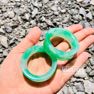 Könnte beinhalten: Zwei Jadeit-Jade-Armreifen mit einem durchscheinenden grün-weißen Marmormuster. Die Armreifen werden in einer Hand gehalten, mit einem felsigen Hintergrund. Der Text "WHITEHOLE" befindet sich in einem weißen Rechteck.