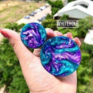 May include: Two round, colourful, resin plugs with a swirling pattern of teal, purple, and white. The plugs are held in a hand.