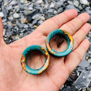 Peut inclure: Une paire d'écarteurs d'oreilles circulaires présentant une base en résine bleu sarcelle lisse avec des motifs tourbillonnants de couleur or métallique et marron incrustés dans le matériau. Le bijou a un centre creux et une finition polie.