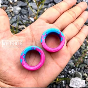 Könnte beinhalten: Zwei kreisförmige Ohr-Dehnstäbe mit einem marmorierten Design in Rosa und Blau. Der Text "WHITEHOLE" ist in einem weißen Feld zu sehen. Die Dehnstäbe werden in einer Hand gehalten.