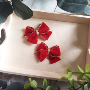 May include: Two red velvet bows with gold trim, resting on a light wooden tray. The bows are a vibrant red, with a gold band in the center and gold trim along the edges. The tray has heart-shaped cutouts on the sides.