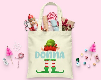 May include: A white canvas tote bag with a green elf hat, red and white striped elf legs, and the name "DONNA" in blue letters. The bag is filled with Christmas-themed items, including candy canes, a nutcracker, and wrapped gifts.