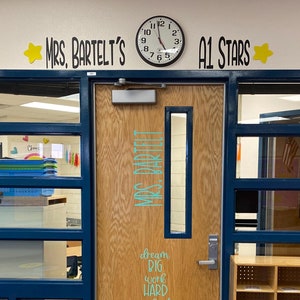 May include: A classroom doorway with a sign that reads "Mrs. Bartelt's" and "A1 Stars" above the door. The door is wood with a glass panel and a sign that reads "Mrs. Bartelt" and "dream big work hard make it happen". There is a bookshelf with cubbies to the right of the door.