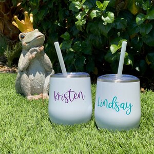 Könnte beinhalten: Zwei weiße isolierte Becher mit Deckeln und Strohhalmen, personalisiert mit den Namen "Kristen" in Lila und "Lindsay" in Türkis. Im Hintergrund eine dekorative Froschstatue mit goldener Krone, auf grünem Gras.