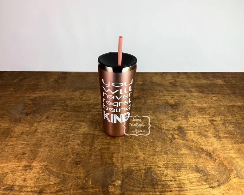 Puede incluir: Un vaso de acero inoxidable color oro rosa con tapa negra y una pajita rosa. El vaso tiene texto blanco que dice "You will never regret being kind".