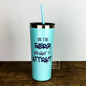 Puede incluir: Un vaso de acero inoxidable azul claro con tapa negra y una pajita. El vaso tiene el texto "Be the energy you want to attract" impreso en &eacute;l.
