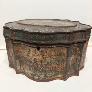 Puede incluir: Una caja de hojalata octogonal antigua con tapa con bisagras. La caja está decorada con un patrón floral verde y dorado alrededor del borde superior y una escena descolorida de caballos y una cerca en los lados. La caja está ligeramente oxidada y muestra signos de antigüedad.
