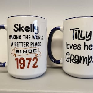 May include: Two white ceramic mugs with blue rims. The mug on the left has the text "Skelly Making the World a Better Place Since 1972". The mug on the right has the text "Tilly loves her Gramps".