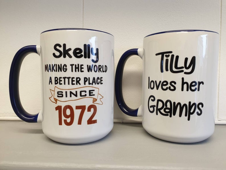 May include: Two white ceramic mugs with blue rims. The mug on the left has the text "Skelly Making the World a Better Place Since 1972". The mug on the right has the text "Tilly loves her Gramps".