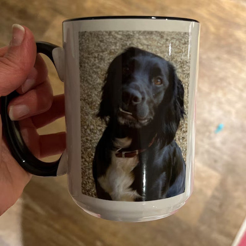 May include: A white ceramic mug with a black rim and handle. The mug features a photo of a black dog with white markings on its chest. The dog is looking directly at the camera.