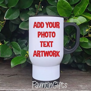 May include: A white travel mug with a black handle and rim. The mug features the red text "ADD YOUR PHOTO TEXT ARTWORK". The mug is set against a backdrop of green foliage and a wooden surface.