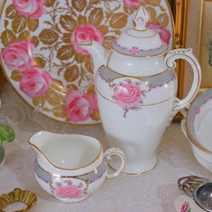 Puede incluir: Un juego de té de porcelana blanca con un patrón de rosas rosas. El juego incluye una tetera, una jarra de crema y un colador de té. La tetera tiene un borde dorado y una tapa con un pomo. La jarra de crema tiene un borde dorado y un asa. El colador de té está hecho de metal y tiene un diseño de malla.