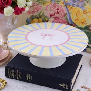 May include: A round ceramic cake stand featuring a white centre with a pink bow illustration, surrounded by alternating pale yellow and light blue radiating stripes. The stand has a gold-toned rim and a solid white pedestal base.