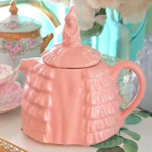 Könnte beinhalten: Eine rosa Keramik-Teekanne mit Rüschendesign und einem hohen, spiralförmigen Knauf auf dem Deckel. Im Hintergrund sind weitere Teetassen und Teekannen zu sehen, darunter eine blaue Teekanne und eine Teetasse mit rosa Blumenmuster.
