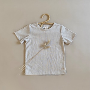May include: A beige ribbed t-shirt with short sleeves and a crew neck, displayed on a wooden hanger. The shirt features a small rainbow logo on the bottom right. A sprig of dried flowers is placed on the shirt. The t-shirt is made of a soft material.