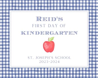May include: A blue and white gingham background with a white rectangle in the center. The rectangle has the text "Reid's First Day of Kindergarten" with a red apple illustration. Below the text is "St. Joseph's School 2023-2024".