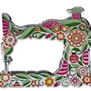 May include: A decorative pin in the shape of a vintage sewing machine. The pin is adorned with a floral design in shades of pink, green, orange, and white. A small ladybug is also featured in the design. The pin is made of metal.