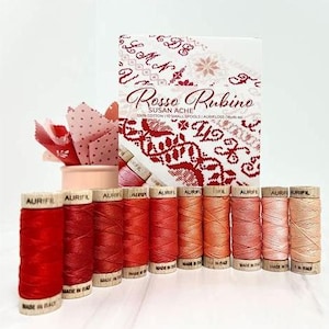 May include: A collection of Aurifil thread spools in various shades of red and orange, arranged in a row. A box labeled "Rosso Rubino" and a small container with fabric are also present. The spools are made in Italy.