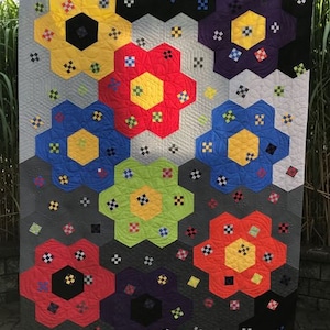 May include: A colorful handmade quilt featuring large, hexagon-shaped floral designs in various colors, including yellow, red, blue, green, and purple. The quilt has a gray background with smaller, square accent patterns. The quilt is displayed outdoors.