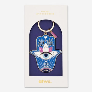 Può includere: Un portachiavi a forma di mano di hamsa in smalto blu con un anello dorato e un design di occhio malvagio bianco. Il portachiavi è confezionato in una scatola bianca con il testo "accessori in smalto" e "atws." stampato su di essa.