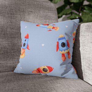 Op de afbeelding: Een lichtblauw kussen met een herhalend patroon van kleurrijke aquarelraketten en kleine witte hartjes. De raketten zijn voornamelijk rood, blauw en geel, met oranje vlammen. Het kussen ligt op een grijze gestoffeerde bank.