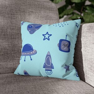 Op de afbeelding: Een vierkant sierkussen met een lichtblauwe achtergrond met een patroon van ruimte-thema illustraties. Het ontwerp bevat een blauw alien gezicht, sterren, een ruimteschip, een raket en een retro tv.