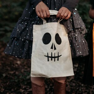 K&ouml;nnte beinhalten: Eine wei&szlig;e Stofftasche mit einem schwarzen Totenkopf-Gesichtsdesign. Die Tasche hat schwarze Henkel und wird von einem Kind getragen, das ein schwarzes Kleid mit silbernen Sternen tr&auml;gt.