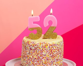 May include: A white cake with pink and gold candles that say "52" on top. The cake is covered in colorful sprinkles.