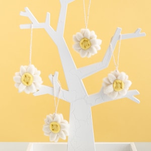 May include: Four white felt flower ornaments with yellow centres hang from a white tree-shaped stand against a yellow background.