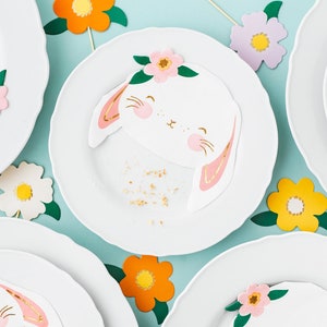 May include: White plates with scalloped edges are decorated with bunny-shaped napkins, featuring pink and gold accents, and floral embellishments. Colorful paper flowers in yellow, orange, and white add to the festive Easter theme.
