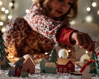 May include: A set of miniature wooden holiday decorations including small houses in red, blue, and green, stylized evergreen trees, a reindeer figure, and small gift boxes, all arranged on a white surface with a dusting of artificial snow.