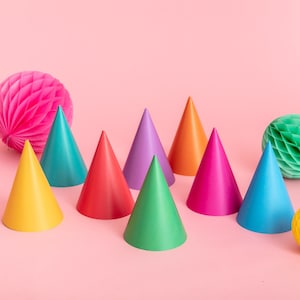 May include: A set of nine colorful party hats in various shades of red, orange, yellow, green, blue, purple, and pink. The hats are arranged in a row on a pink background.