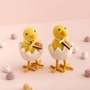 May include: Two yellow felt chicks in white egg shells with yellow ribbon bows and personalised wooden name tags. The chick on the left has a name tag that says "Ella" and the chick on the right has a name tag that says "Harry".