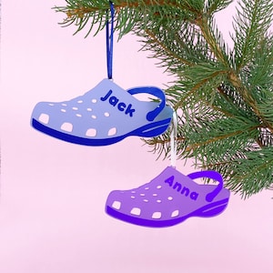 May include: Two blue and purple personalised Christmas decorations shaped like a clog shoe. The blue ornament has the name "Jack" on it and the purple ornament has the name "Anna" on it.