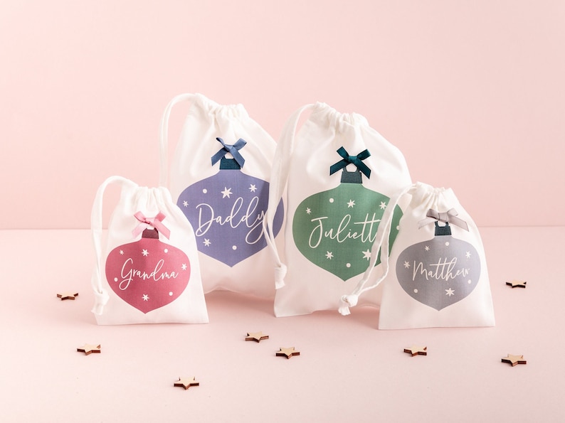 K&ouml;nnte beinhalten: Vier wei&szlig;e Geschenkbeutel mit Kordelzug und Christbaumschmuck-Designs. Jeder Beutel hat eine andersfarbige Verzierung mit einem Namen: Grandma, Daddy, Juliett und Matthew. Die Beutel haben eine Schleife oben und sind auf rosa Hintergrund.