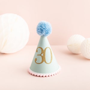 May include: A light blue party hat with a white pom-pom on top and a gold glitter number "30" on the front. The hat has a pink and white trim around the bottom.
