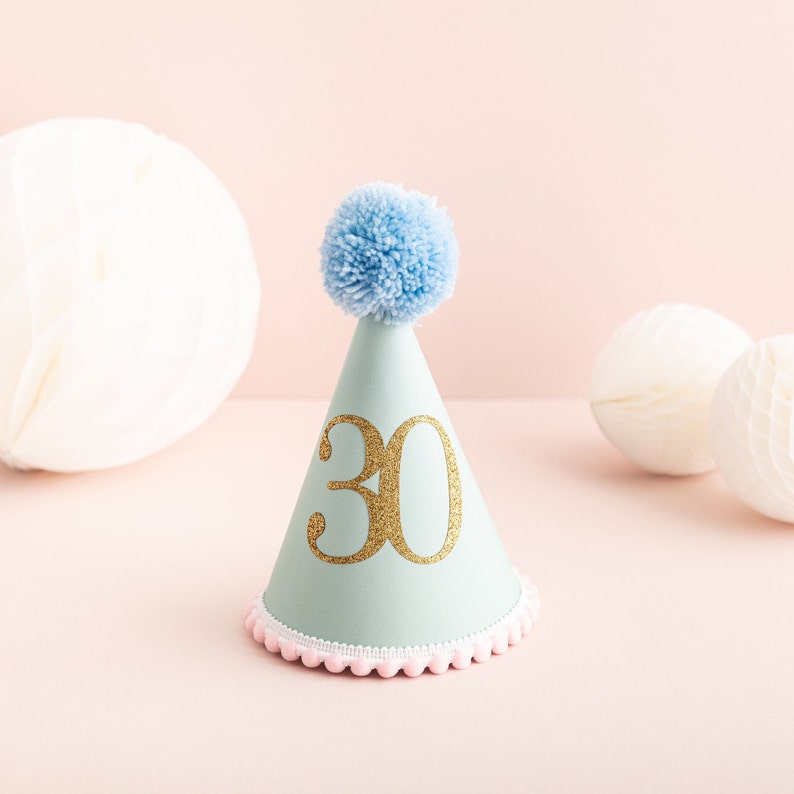 May include: A light blue party hat with a white pom-pom on top and a gold glitter number "30" on the front. The hat has a pink and white trim around the bottom.