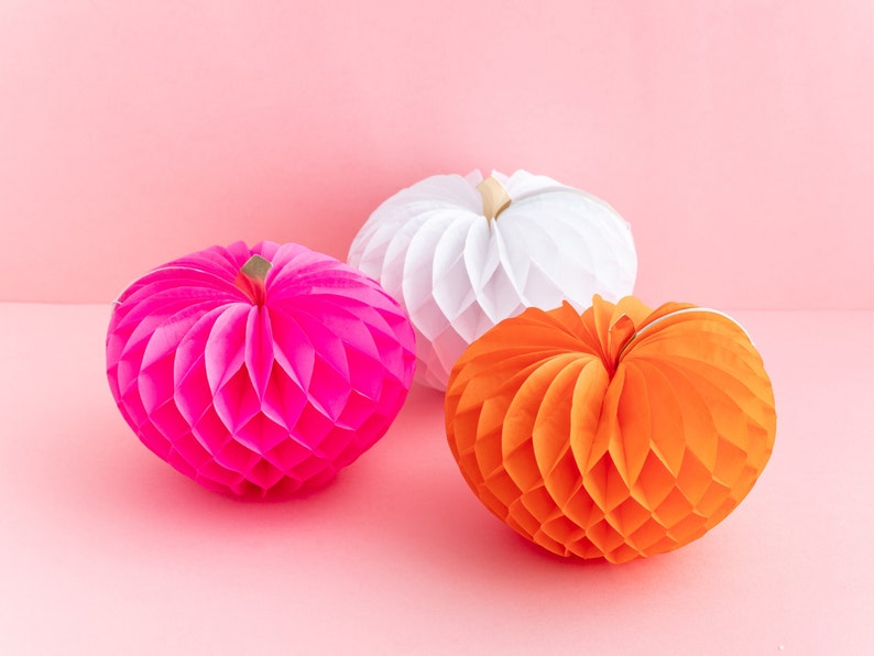 May include: Three honeycomb paper pumpkins in pink, white, and orange. The pumpkins are on a pink background.