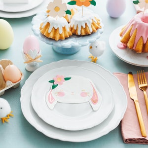 May include: An Easter-themed table setting with pastel-colored egg candles, cakes with floral toppers, and bunny-faced plates. Gold cutlery and decorative chicks add to the festive atmosphere. The table is set on a light blue tablecloth.