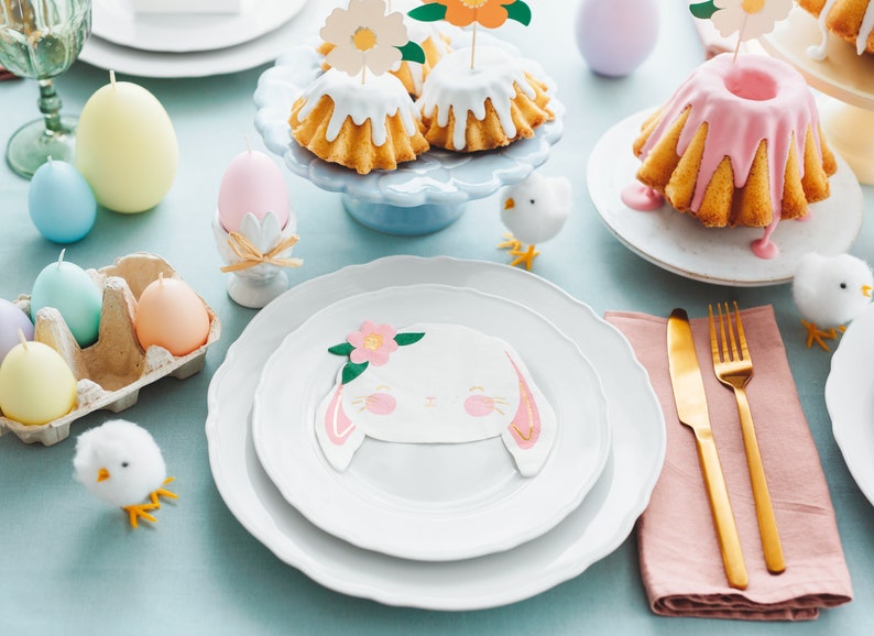 May include: An Easter-themed table setting with pastel-colored egg candles, cakes with floral toppers, and bunny-faced plates. Gold cutlery and decorative chicks add to the festive atmosphere. The table is set on a light blue tablecloth.