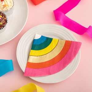 May include: A rainbow-colored paper napkin with a gold stripe, folded into a triangle shape, on a white plate. The napkin is on a pink surface with colorful ribbons.