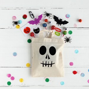 K&ouml;nnte beinhalten: Eine wei&szlig;e Stofftasche mit einem schwarzen Totenkopf- und Knochenmotiv. Die Tasche ist gef&uuml;llt mit Halloween-S&uuml;&szlig;igkeiten, darunter Bonbons, Lutscher und Aufkleber. Die Tasche ist von buntem Konfetti und anderen Halloween-Dekorationen umgeben.