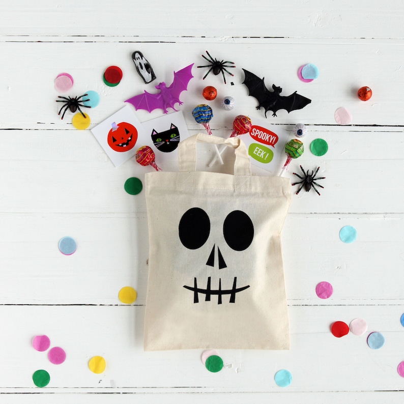 K&ouml;nnte beinhalten: Eine wei&szlig;e Stofftasche mit einem schwarzen Totenkopf- und Knochenmotiv. Die Tasche ist gef&uuml;llt mit Halloween-S&uuml;&szlig;igkeiten, darunter Bonbons, Lutscher und Aufkleber. Die Tasche ist von buntem Konfetti und anderen Halloween-Dekorationen umgeben.
