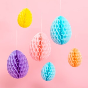 May include: Six pastel-colored honeycomb paper Easter eggs hanging from strings against a pink background. The eggs are in shades of yellow, blue, pink, purple, and orange.