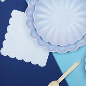 May include: Two lavender paper plates with scalloped edges, a white paper napkin with scalloped edges, and a wooden spoon on a blue and light blue background. The napkin has the text "Made with Love" printed on it.